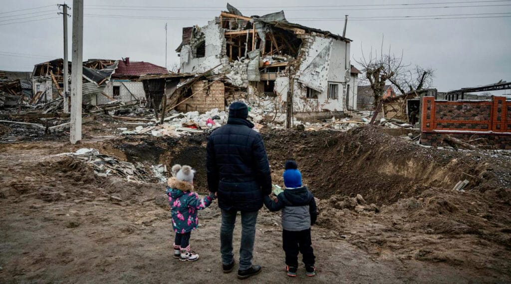 A Ukrainian father and two young children stand holding hands, looking toward their severely damaged home, reduced to rubble and debris by war.